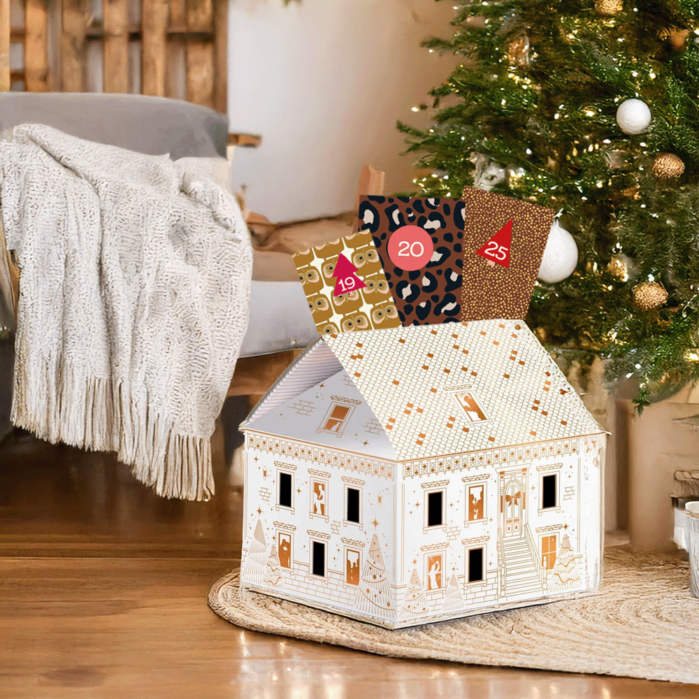 Een witte cadeaudoos in de vorm van een huis, en er steken wat cadeauzakjes uit. Op de achtergrond staat een kerstboom